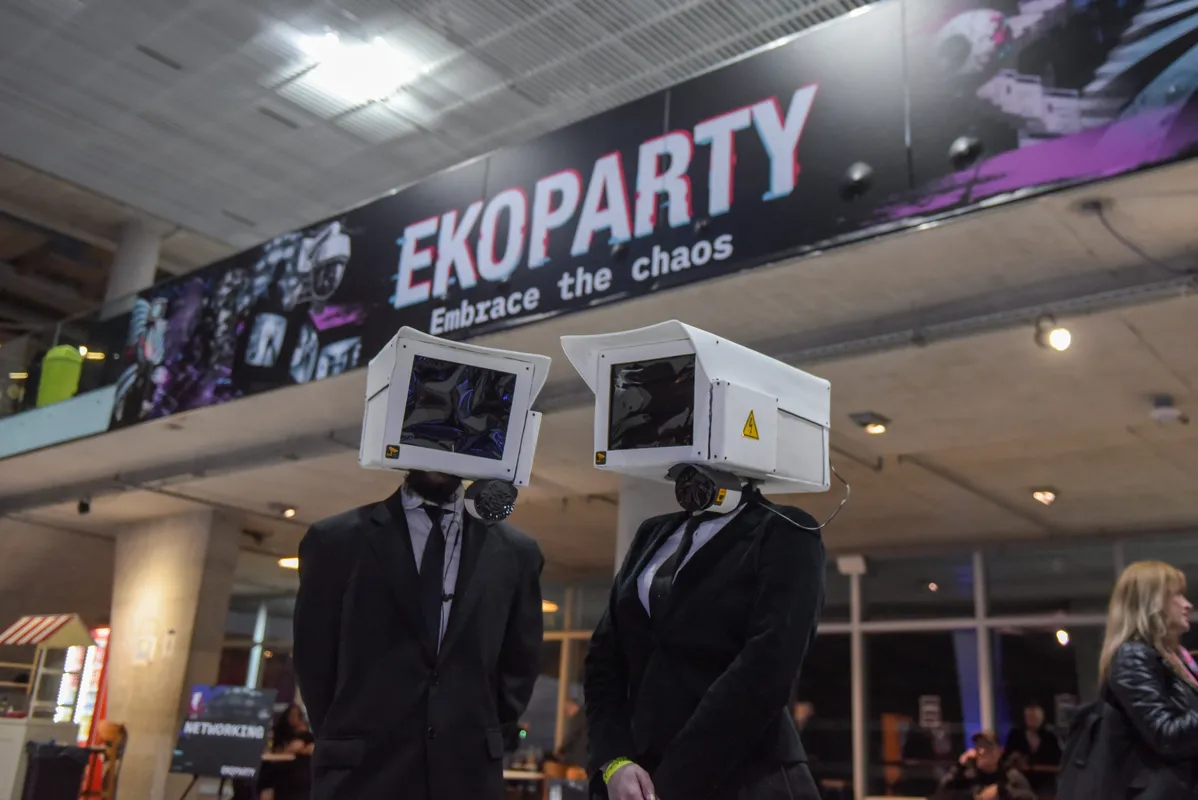 Two people in black suits wearing surveillance-camera boxes as heads, under a banner that reads "Ekoparty: Embrace the chaos"