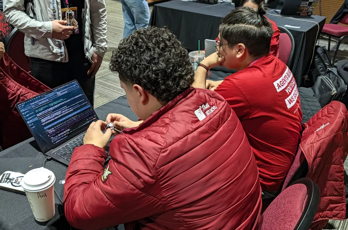 Two players in red jackets at a CTF table, one holding a small device next to a laptop with a terminal open