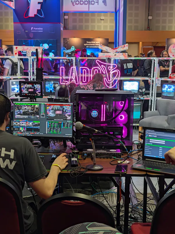 The Lado B streaming desk: monitors with the live switcher, a camera, a pink-lit PC and a "LADO B" neon sign, with the conference floor behind